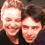 Two people in a close-up pose, one with light brown hair and the other with dark hair, smiling at each other against a dark background.