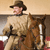 A pixelated figure in a brown hat and light-colored coat stands beside a brown horse, holding reins, against a muted background.