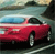 A red sedan with a light blue roof is positioned on a road, viewed from a low angle with a blurred background of green and brown tones.
