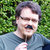 Pixelated static image of a person with short dark hair wearing a light blue shirt against a green foliage background.
