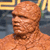 Pixelated close-up of a bald figure with light brown skin tone, wearing a dark brown collar, against a blurred background with yellow and blue elements.