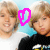 Two young people with light brown hair stand against a blue background; one wears white, the other black. A pink heart symbol animates in three distinct states.