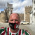 A pixelated image shows a person with a bald head wearing a dark mask, red and green striped clothing, and a white collar, standing before a blurred background of stone structures under a light sky.