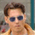 A person with brown hair and blue sunglasses wears a yellow jacket over a light shirt against a blurred background.