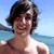 Pixelated image of a person with dark hair wearing a white top, standing against a blue sky and water background.