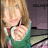 Pixelated image of a person with long blonde hair wearing a green and white top, holding an object near their face against a dark background.