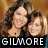 Two women with long brown hair pose closely together against a warm-toned background, one wearing a dark top and the other in blue.