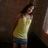 A pixelated figure in a yellow sleeveless top and blue shorts stands against a dark brown background with partial light-colored vertical element on the right.