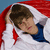 A person with brown hair lies under a red and white blanket, wearing a blue shirt, with eyes closed and a slight smile.