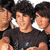 Three individuals with dark hair and light skin wear black shirts against a white background, positioned closely together in a group pose.