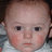 Pixelated close-up of a child's face with light skin, dark eyes, and rosy cheeks, wearing a light-colored garment with a blue patterned background.