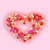 A heart-shaped arrangement of multicolored pixelated flowers on a light pink background with a soft gradient effect.