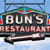 Pixelated sign for Bun's Restaurant featuring bold text, a baseball bat graphic, and a blue sky background with structural elements.