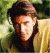 Pixelated image of a person with brown hair wearing a light-colored jacket over dark clothing against green foliage background.