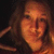 A person with long brown hair in a dark setting, wearing an orange top and making hand gestures near their head.