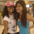 Two women pose together in a shopping mall; one wears a pink cap and white shirt, the other a blue top, with background lights and blurred storefronts.