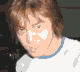 Two side-by-side pixelated frames show a person with light brown hair wearing a white shirt with blue collar, first looking down then making a peace sign with right hand.