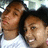 Two people with brown hair and light skin pose closely together, one in a white shirt, against a blurred background with light and dark tones.