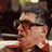 Pixelated static image of a person's face in profile with dark hair, glasses, and red clothing against a blurred background.