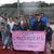 Group of people standing behind a pink sign with indistinct text, set against a blurred background.