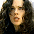 Pixelated close-up of a person with long dark curly hair, light skin, and a neutral expression, wearing dark clothing against a dark background.