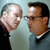 Two men face each other in close proximity against a light blue background, one with a bald head and the other with dark hair and glasses.
