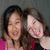 Two people with dark hair pose closely together, one in a pink top and the other in red and black clothing against a gray background.