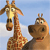 Two pixelated cartoon animals stand against a light blue sky with white clouds; a giraffe with a long neck and patterned coat is on the left, and a smaller brown animal with a wide mouth is on the right.