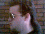 A person with dark hair and glasses appears in two side-by-side frames against a brick wall background, showing slight head movement.