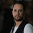 Pixelated static image of a person with short dark hair wearing a black vest over a white shirt against a dark background.