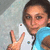 A person in a light blue sleeveless top holds up a white smartphone with two small colored dots on its back, making a peace sign with their right hand against a textured gray background.