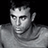 Static grayscale pixel art of a person's upper body and face with dark hair, visible facial features including eyes, nose, mouth, and ear, wearing a light-colored shirt against a black background.