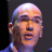 Pixelated close-up of a bald person with light skin tone wearing blue clothing against dark background.