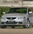 Static image of a silver sedan with black accents parked on a paved surface, green foliage in the background, and visible headlights and grille.