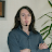Pixelated image of a person with long dark hair wearing a dark blue shirt, standing indoors with a framed item on the wall and green foliage visible.