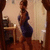 A person in a blue dress stands in a room with wooden flooring and a table, captured in a static image with no visible motion.