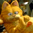 A pixelated orange tabby cat with green eyes and pink inner ears, positioned slightly left of center against a blurred background of indistinct shapes and muted colors.