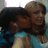 Two figures in close embrace, one with dark hair and blue clothing, the other with light brown hair and a beige top against a blurred background.