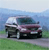 Pixelated image of a dark-colored sedan with a light roof on a road, set against a blurred background of green grass and blue sky.