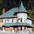 Pixelated static image of a two-story house with light blue roof, white walls, and dark brown trim, set against a dark green background.
