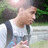 Pixelated image of a person with light brown hair wearing a white sleeveless top and dark straps, standing outdoors near green foliage.