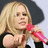 A blonde woman holding a pink microphone, wearing a black and white top, with a dark background and light gray foreground.