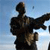 Pixelated figure in dark clothing with arms extended against a light blue sky background.