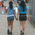 Two women in blue tops and dark shorts walk away from the viewer on a sidewalk, with one woman's right arm raised.