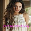 A woman with long wavy brown hair wearing a white sleeveless lace top, standing indoors with a blurred background and pink text overlay.