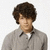 Pixelated image of a person with dark curly hair wearing a gray collared shirt over a white undershirt against a plain light background.