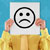 A pixelated image shows a person holding up a white card with a sad face emoji against a blue background.