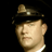 Static pixelated image of a person wearing a dark cap with a light emblem and white collar against black background.