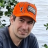 A pixelated portrait of a person wearing an orange cap with gray accents and dark clothing, set against a blurred background with green foliage.