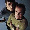 Two men in Star Trek uniforms pose closely together, one in a gray uniform with a black collar, the other in a gold uniform with a black collar and a triangular insignia.